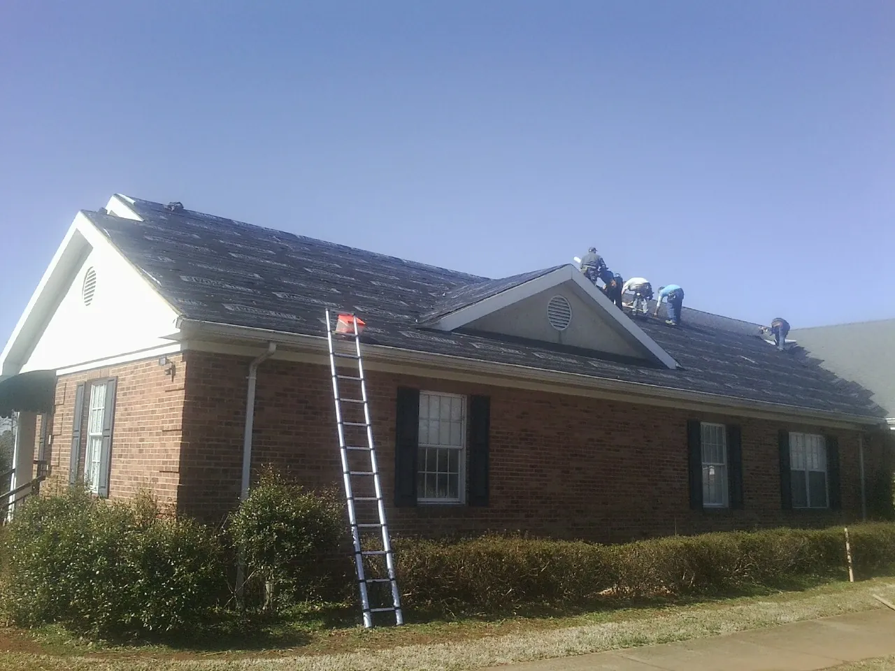 Crew tearing shingles off a brick ranch roof with a ladder set — Emergency Roof Repair work by our crew in Rush Center