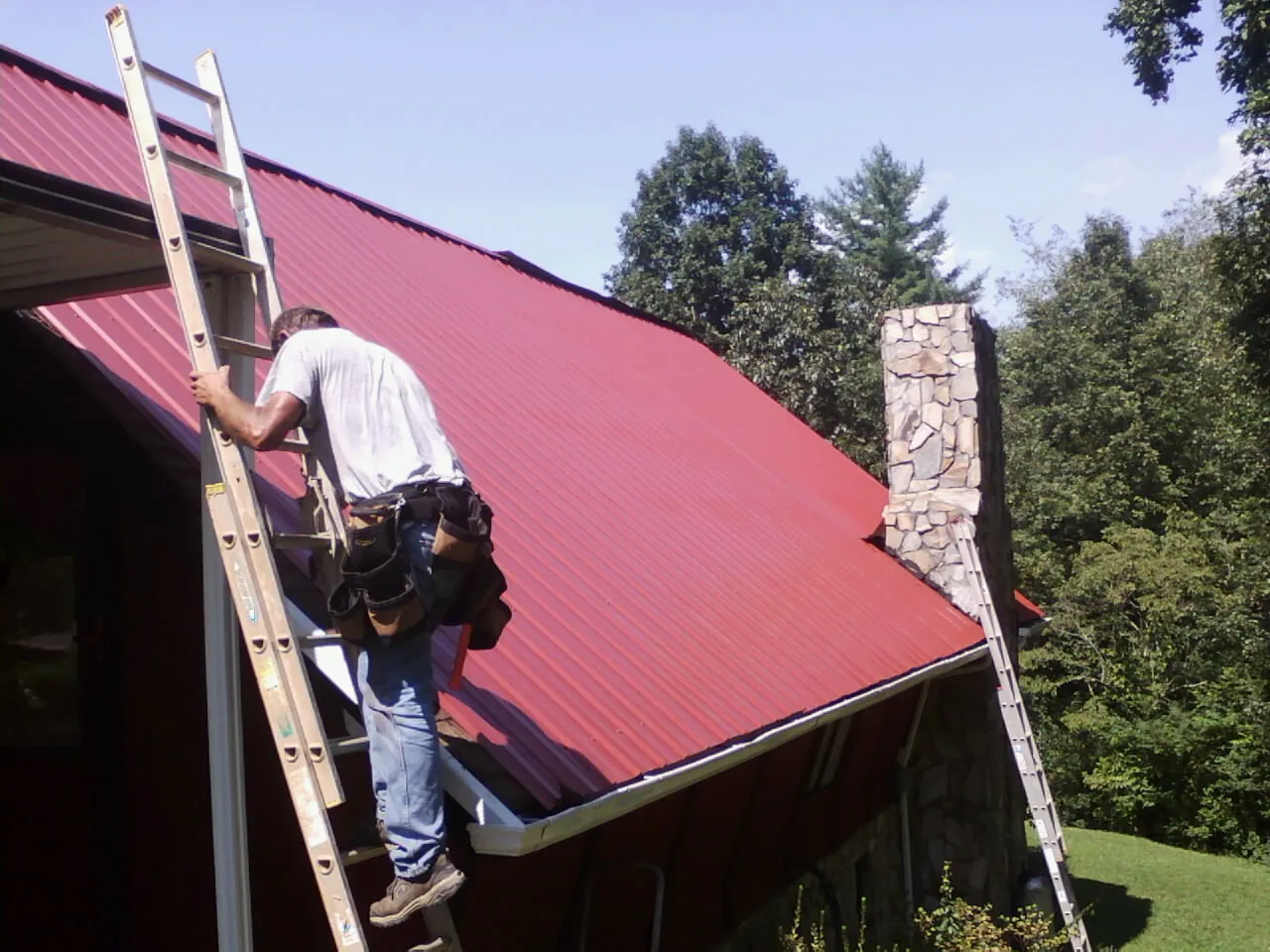 Roofer on a ladder beside a red metal roof and stone chimney — Soffit & Fascia Repair by Roofing Brothers in Rush Center